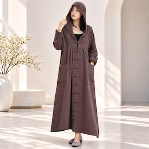 May include: A long-sleeved, floor-length brown hooded coat featuring a front closure with multiple fabric toggle buttons. The garment includes two large patch pockets on the front and is constructed from a textured, lightweight fabric.