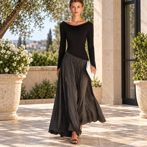 May include: A floor-length dress featuring a black long-sleeved bodice with a draped cowl neckline and a contrasting charcoal grey pleated skirt. The garment is paired with a small white clutch and black open-toe sandals, creating a cohesive look.