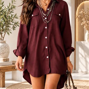 May include: A long-sleeved, button-down shirt in a deep burgundy color. The shirt features a classic collar, two chest pockets with buttoned flaps, and a high-low hemline. The fabric appears smooth and lightweight, with white buttons down the front.