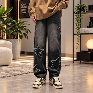 May include: Loose-fitting black denim jeans featuring a distressed star pattern with frayed edges. The jeans are paired with a tan long-sleeve shirt and black and white patterned sneakers. The denim has a faded wash and a wide-leg silhouette.