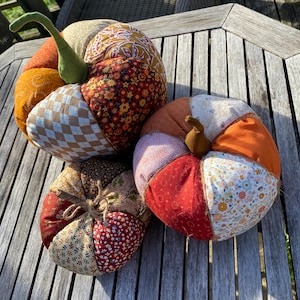 Puede incluir: Tres calabazas de tela rellenas de varios tamaños, con diseños de patchwork con patrones florales, a cuadros y sólidos en tonos naranja, rojo, marrón y crema. Cada calabaza tiene un tallo de tela o cordel sujeto en la parte superior.