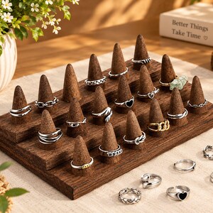May include: A tiered wooden ring display stand featuring multiple cone-shaped holders. Several silver-toned metal rings with various designs, including chains, hearts, and a green stone, are arranged on the cones and on the surface.