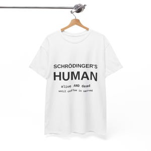 May include: A white short-sleeved t-shirt featuring black text that reads SCHRÖDINGER'S HUMAN, followed by the text alive AND dead, and finally the text until coffee is served. The garment is displayed on a wooden hanger against a plain background.