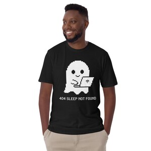 Könnte beinhalten: Ein schwarzes Kurzarm-T-Shirt mit einer weißen, verpixelten Grafik eines Geistes, der einen Laptop benutzt. Unter dem Geist steht der Text 404 SLEEP NOT FOUND in einer weißen, verpixelten Schriftart. Das Shirt hat einen Rundhalsausschnitt.