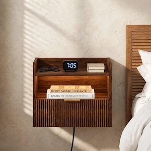 May include: A wall-mounted dark wood nightstand featuring a fluted drawer front with a gold handle, an open shelf with integrated warm lighting, a digital clock displaying 7:05, black eyeglasses, and two books titled War and Peace and Chanel.