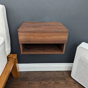 May include: A wall-mounted floating nightstand featuring a dark brown wood grain finish. The unit includes a single horizontal drawer at the top and an open storage shelf directly underneath, providing a compact and functional bedside storage solution.
