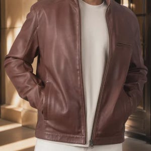 May include: A brown leather jacket featuring a stand-up collar, a full-length front zipper, and two side pockets. The jacket is worn over a plain light-colored t-shirt and light-colored pants, showcasing a smooth, solid-colored finish.