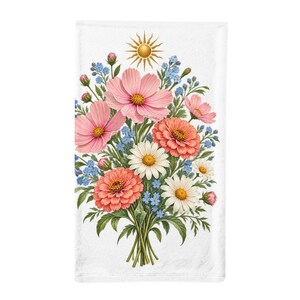 May include: A white rectangular towel featuring a central floral bouquet of pink cosmos, orange zinnias, white daisies, and small blue forget-me-nots. A golden sun icon is positioned above the flowers, which have green stems and leaves.