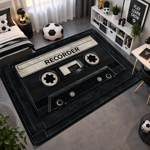 Könnte beinhalten: Ein rechteckiger Teppich mit einer großen, detaillierten Grafik einer schwarz-weißen Kassette. Das Design umfasst zwei kreisförmige Spulen, ein Sichtfenster in der Mitte und das Wort RECORDER, das in fetten schwarzen Großbuchstaben oben steht.