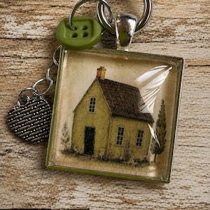May include: A square pendant keychain featuring a printed illustration of a yellow house with a dark roof and chimney. The pendant is attached to a silver-toned metal ring, a green four-hole button, and a small heart-shaped charm with a textured pattern.