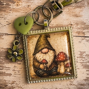 May include: A square metal keychain featuring a gnome illustration with a long white beard and a brown hat, sitting next to red and white mushrooms. The keychain includes a silver lobster clasp, a light green heart-shaped button, and a flower charm.