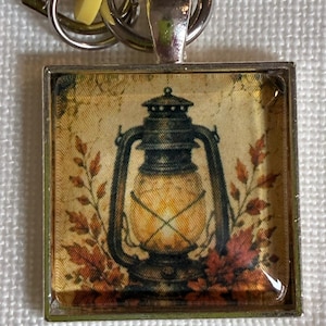May include: A square silver-toned pendant keyring featuring a glass-covered image of a vintage lantern glowing with warm light, surrounded by orange and brown autumn leaves against a textured, aged cream-coloured background with a metal ring attachment.