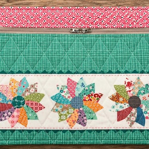 May include: A rectangular quilted fabric pouch featuring a top section with a pink and white geometric pattern, a middle section in green with a diamond grid, and a bottom white section displaying three colourful floral-shaped fabric appliqués.