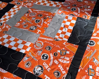 May include: A quilted blanket featuring a patchwork design with orange, black, white, and gray squares. The orange squares are printed with the text "Tennessee", "Vols", and various team-related graphics. The blanket has a black border.