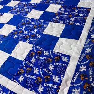 May include: A blue and white quilt with a repeating pattern. The quilt features squares of blue fabric with the text "Kentucky Wildcats" and a wildcat logo, alternating with white squares. The quilt is machine-stitched.