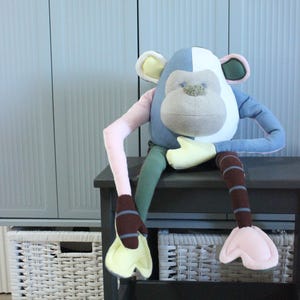 Può includere: Un peluche a forma di scimmia con un design a blocchi di colore composto da pannelli in azzurro, bianco, rosa pallido, verde scuro e marrone. Il peluche ha il viso grigio chiaro, mani e piedi gialli e gambe marroni a righe con piedi rosa.