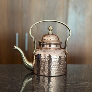 May include: A hammered copper tea kettle featuring a polished brass handle, a matching brass spout, and a domed lid topped with a small brass finial. The body of the kettle has a textured, dimpled surface that reflects light across its metallic finish.