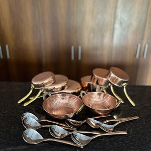 以下が含まれることがあります： 銅色の調理器具セット。側面ハンドル付きの丸いハンマー仕上げのボウル2個、蓋と真鍮色のハンドルが付いた小さな鍋数個、そして手前に配置された反射する金属表面を持つ複数のマッチングスプーンが含まれています。