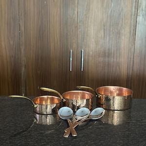 May include: Three hammered copper saucepans of varying sizes, each featuring a polished brass handle. Accompanying the pans are three silver-toned metal spoons arranged in a cluster in front of the cookware, reflecting the surrounding light.