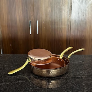 May include: Two round copper pans with a hammered exterior finish and polished gold-tone metal handles. One pan is placed inside the other, with the smaller pan inverted on top, showcasing the smooth copper base and the textured side walls.