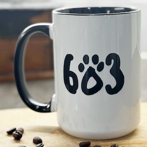 May include: White ceramic mug with a black handle and rim, featuring the number "683" in a paw print design. The mug sits on a wooden cutting board with scattered coffee beans. A vintage coffee grinder is in the background.