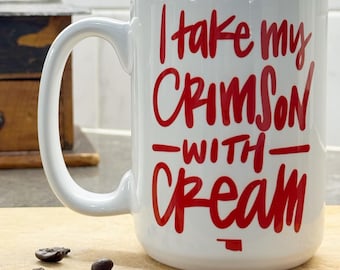 May include: White ceramic mug with the text "I take my CRIMSON with CREAM" in red lettering. The mug sits on a wooden cutting board with coffee beans. A vintage coffee grinder is in the background.