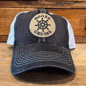 May include: Black and white trucker hat with a curved brim. The hat features a leather patch with the words "BOOZE CRUISE" and a ship's wheel design. The hat is sitting on a wooden surface.