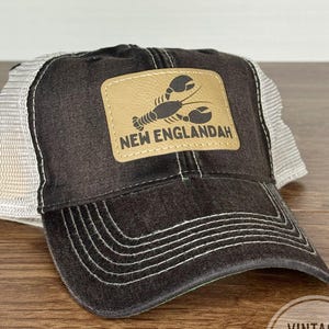 May include: A black and gray trucker hat with a tan leather patch featuring a lobster graphic and the text "NEW ENGLANDAH." The hat has a mesh back and a curved brim. The hat is sitting on a wooden surface.