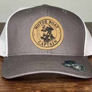 May include: A gray trucker hat with a white mesh back and a curved brim. The hat features a circular patch with the words "MOTOR BOAT CAPTAIN" around an illustration of a bearded man smoking a pipe.