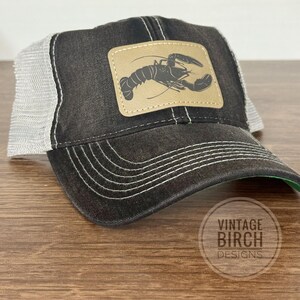 May include: A black trucker hat with a light grey mesh back and a brown leather patch featuring a lobster design. The hat has white stitching and the text "VINTAGE BIRCH DESIGNS" is printed on the bottom right.