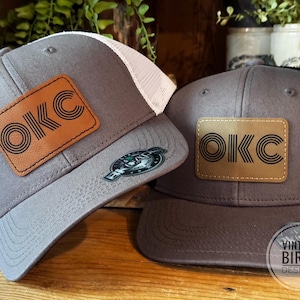 May include: Two gray trucker hats with the letters OKC on a brown leather patch. One hat has a white mesh back, the other has a black mesh back. The hats are on a wooden surface. The hats are from Vintage Birch Designs.