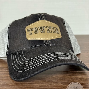 May include: Black and gray trucker hat with a white mesh back. The front panel features a tan leather patch with the word "TOWNIE" in capital letters. The hat has a curved brim and white stitching details.