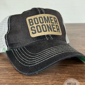 May include: Black trucker hat with a white mesh back and a curved brim. The hat features a tan leather patch with the words "BOOMER SOONER" in bold, block letters. The hat has white stitching and a green underside.