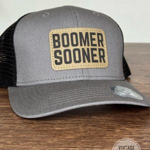 May include: Gray trucker hat with a black mesh back and a curved brim. The hat features a tan leather patch with the words "BOOMER SOONER" in bold, black letters. The hat has a snap closure and a small tag on the brim.
