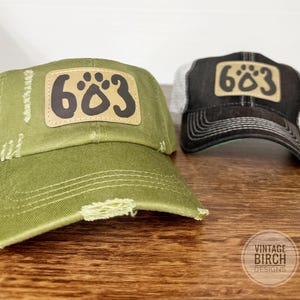 May include: Two baseball caps with the number "683" on a patch. One cap is olive green with distressed details, and the other is black with a white mesh back. The caps are on a wooden surface.