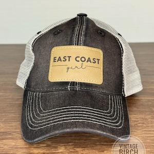 May include: Black trucker hat with a distressed look, featuring a tan leather patch with the words "EAST COAST girl" in a script font. The hat has a white mesh back and a curved brim with white stitching.