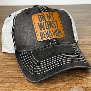 May include: Black and gray baseball cap with a white mesh back. The cap features a brown leather patch with the phrase "ON MY WORST BEHAVIOR" in black lettering. White stitching accents the brim and seams.