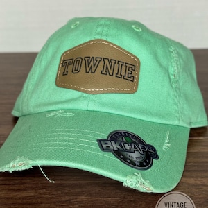 May include: A distressed mint green baseball cap with a brown leather patch that reads "TOWNIE". The cap has a curved brim and a faded appearance. A sticker with the text "BK CAPS" is visible on the side.