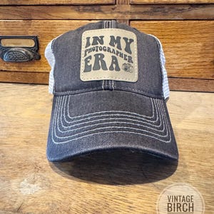 May include: A denim-style baseball cap with a white mesh back. The front features a tan square patch with the words "IN MY PHOTOGRAPHER ERA" and a camera icon. White stitching accents the brim and the cap's front.