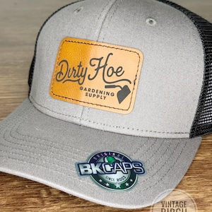 May include: A gray and black trucker hat with a leather patch that reads "Dirty Hoe Gardening Supply." The hat has a curved brim and a mesh back. The hat also has a "BK Caps" sticker and a "Vintage Birch Designs" logo.