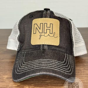 May include: A black trucker hat with a white mesh back and a curved brim. The hat features a tan leather patch with the text "NH girl" in a handwritten font. White stitching accents the brim and the hat's seams.