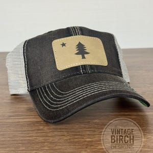 May include: A black trucker hat with a white mesh back and a curved brim. The hat features a tan patch with a black tree and star design. The text "VINTAGE BIRCH DESIGNS" is visible on the bottom right.