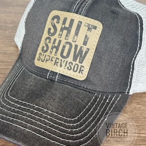 May include: A distressed black and white trucker hat with a tan leather patch. The patch features the words "SHIT SHOW SUPERVISOR" in a distressed font. The hat has a curved brim and a mesh back.