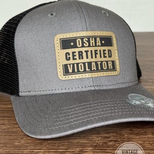 May include: A gray trucker hat with a black mesh back and a curved brim. The hat features a rectangular patch with the text "OSHA CERTIFIED VIOLATOR" in black letters on a tan background. The hat is a vintage design.