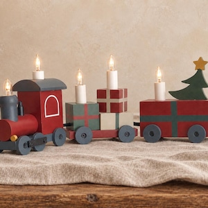 Op de afbeelding: Een decoratieve houten treinlamp met een rode en donkergrijze locomotief, een middenwagon met gestapelde cadeauverpakkingen en een achterwagon met een groene kerstboom. De trein bevat vier elektrische kaarslampjes en een gele ster als piek.