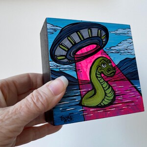 May include: A hand-painted wooden block with a green snake being abducted by a flying saucer. The saucer is casting a pink beam of light on the snake. The background is a blue sky with clouds and mountains.