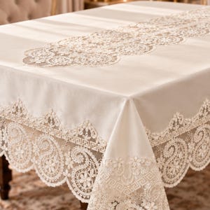 May include: A cream-colored rectangular tablecloth featuring an intricate lace border with floral motifs and a geometric Greek key pattern. A matching lace runner with circular floral designs is centered along the top of the smooth fabric surface.