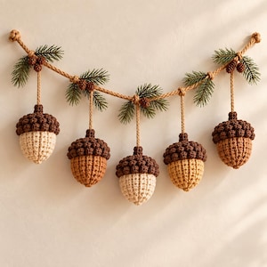 May include: A decorative garland featuring five crocheted acorns in shades of cream, tan, and brown, suspended from a thick twisted rope. The rope is adorned with green pine needle sprigs and small brown pinecones at each hanging point.