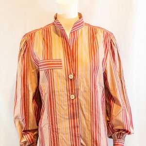 May include: A long-sleeved, striped button-up shirt with a mandarin collar. The shirt is made of a lightweight fabric and has a relaxed fit. The colors are a mix of red, yellow, and purple.