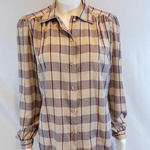 May include: A button-down shirt with a plaid pattern in beige, gray, and burgundy. The long-sleeved shirt has a collared neckline and decorative buttons. The fabric has a subtle dotted texture. The shirt is displayed on a mannequin.